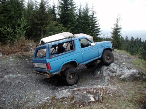 1985 Ford Bronco - A blue first-generation Ford Bronco is navigating a rocky, off-road trail with a second vehicle partially visible behind it. The Bronco is…