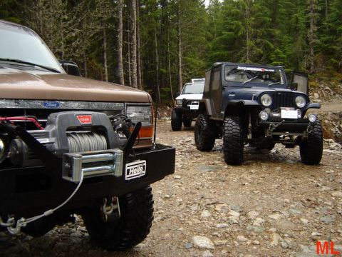 1985 Ford Bronco - A line of off-road vehicles, including a brown Ford Bronco with a winch and a dark blue Jeep, are parked on a dirt trail in a forest.