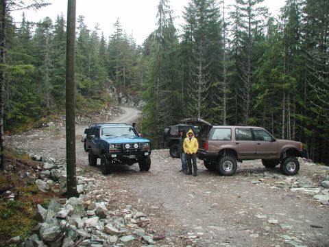 1985 Ford Bronco - Two modified SUVs are parked on a gravel road in a forested area. The blue SUV is in the foreground, and a brown SUV with its rear hatch op…