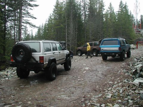 1985 Ford Bronco - A group of off-road vehicles are parked on a wet, rocky trail surrounded by a dense forest. The lead vehicle is a silver Jeep Cherokee with…