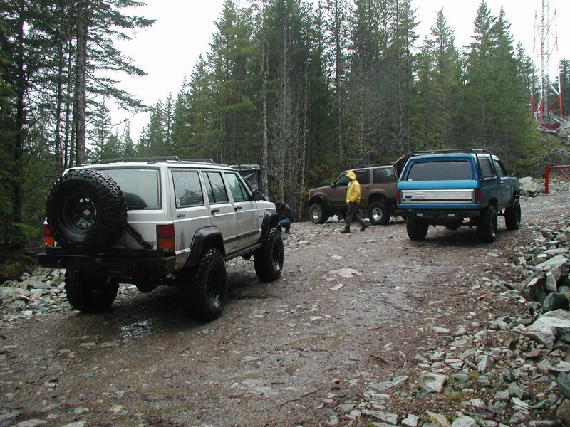 Silver Jeep Cherokee, brown SUV, and blue Ford Bronco on a wet forest trail.