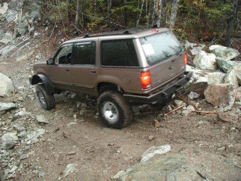 1985 Ford Bronco - A lifted, brown SUV with aggressive tires navigates a rocky, muddy trail in a wooded area. The vehicle is angled to the right, showing its…