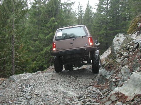 1985 Ford Bronco - A brown SUV is navigating a rocky, off-road trail surrounded by dense evergreen trees. The vehicle is tilted to the right as it climbs over…