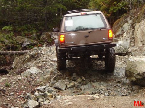 1985 Ford Bronco - A brown Ford Explorer is navigating a rocky, off-road trail. The rear of the SUV is visible as it climbs over large stones and uneven terra…