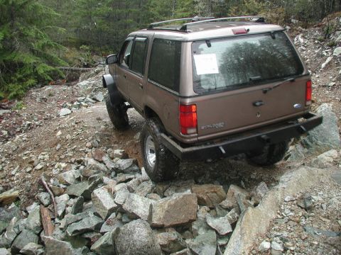1985 Ford Bronco - A lifted, brown Ford Explorer is navigating a rocky, off-road trail with large boulders and trees visible in the background.
