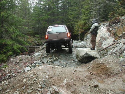 1985 Ford Bronco - A lifted brown SUV navigates a rocky, off-road trail with a person standing on a boulder watching. The vehicle is positioned on a steep inc…