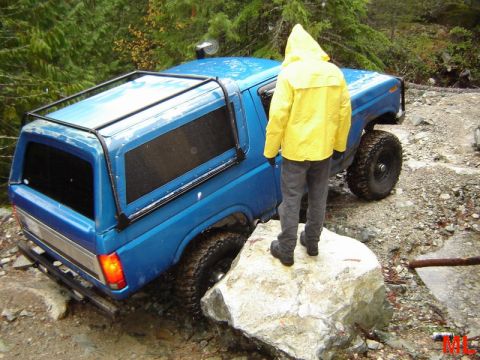 1985 Ford Bronco - A blue Ford Bronco with a camper shell is navigating a rocky, wooded trail. A person wearing a yellow raincoat stands on a large rock next…