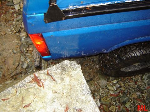 1985 Ford Bronco - The rear corner of a blue pickup truck is shown with its taillight illuminated, positioned next to a large rock and rocky terrain.
