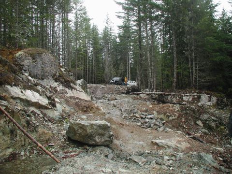 1985 Ford Bronco - Two off-road vehicles, a black SUV and a white SUV, are parked on a rocky, wooded trail. The terrain is rough with large boulders and trees…