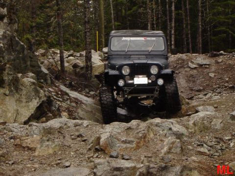 1985 Ford Bronco - A dark colored Jeep is navigating a rocky, off-road trail in a forest. The vehicle has large, knobby tires and appears to be climbing over…