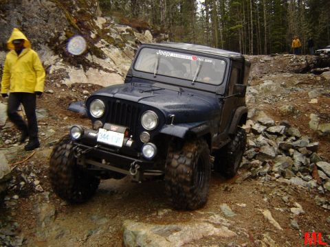 1985 Ford Bronco - A dark blue Jeep CJ is navigating a rocky, off-road trail. A person in a yellow raincoat walks alongside the Jeep.