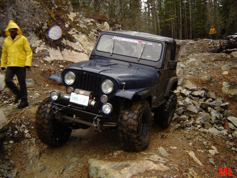 Dark blue Jeep CJ off-roading on a rocky trail with a person in a yellow raincoat.