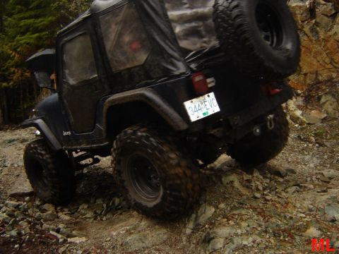 1985 Ford Bronco - A black Jeep Wrangler is navigating a rocky, off-road trail. The vehicle is angled upwards, with its large tires gripping the uneven terrai…