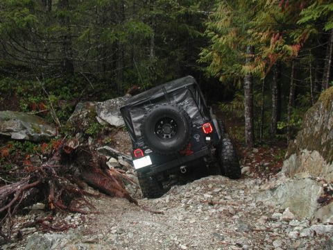 1985 Ford Bronco - A dark-colored Jeep Wrangler is navigating a steep, rocky off-road trail in a dense forest. The vehicle is tilted significantly to the righ…