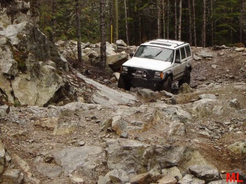 1985 Ford Bronco - A white Jeep Cherokee is navigating a rocky, off-road trail with dense trees in the background. The vehicle is angled towards the viewer's…