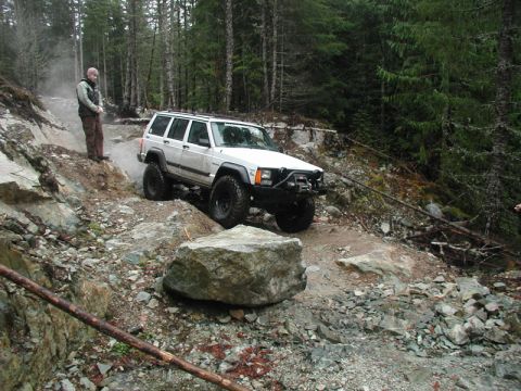 1985 Ford Bronco - A white Jeep Cherokee XJ navigates a rocky, off-road trail with a person standing on the side watching. The vehicle is positioned on a stee…