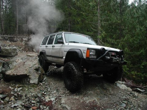 1985 Ford Bronco - A white Jeep Cherokee is navigating a rocky, off-road trail with trees in the background. Smoke or steam is rising from the vehicle.
