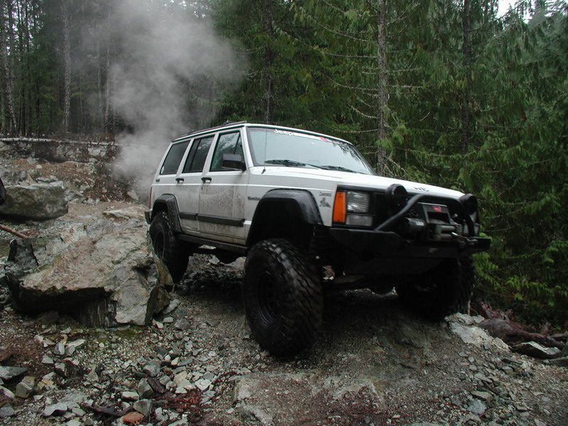 White Jeep Cherokee off-roading on a rocky trail with smoke rising.