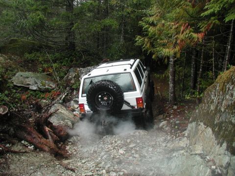 1985 Ford Bronco - A white Jeep Cherokee is navigating a rocky, wooded trail, kicking up dust and smoke from its tires. The vehicle is viewed from the rear as…