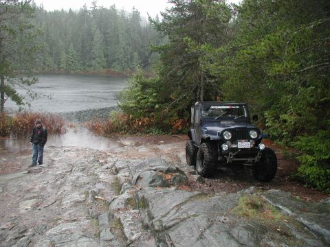 1985 Ford Bronco - A dark blue Jeep CJ-7 with large off-road tires is parked on a rocky, wet trail next to a lake. A person in a black jacket stands on the ro…