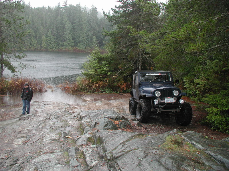 Dark blue Jeep CJ-7 on a rocky trail by a lake in the rain.
