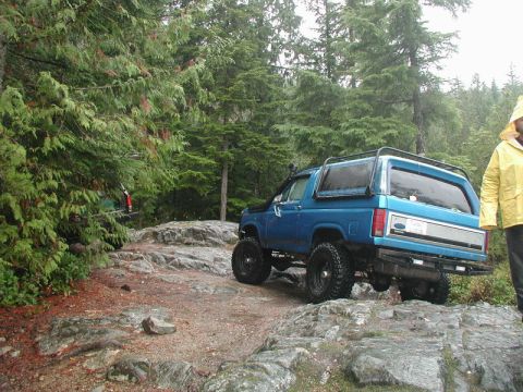 1985 Ford Bronco - A blue Ford Bronco is navigating a rocky, off-road trail surrounded by dense evergreen trees. A person in a yellow raincoat stands to the r…
