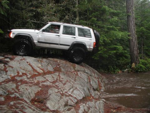 1985 Ford Bronco - A white Jeep Cherokee is navigating a rocky, off-road trail with water on the right side of the frame. The vehicle is positioned with its f…