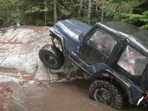1985 Ford Bronco - A dark blue Jeep Wrangler is navigating a muddy water obstacle on a rocky terrain, with its tires deep in the water and one tire on a large…
