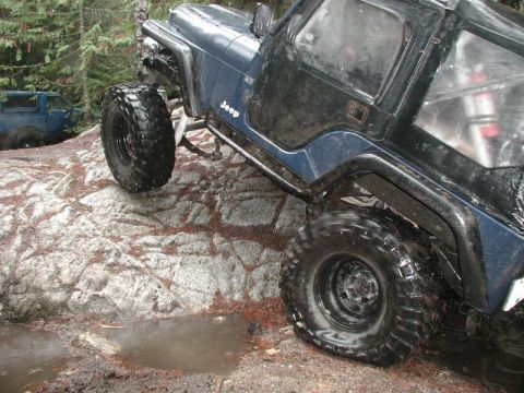 1985 Ford Bronco - A dark blue Jeep is shown from the side, navigating over a large, wet rock formation in a wooded area. The Jeep has large, knobby tires and…