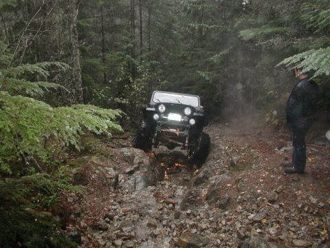 1985 Ford Bronco - A black Jeep Wrangler is navigating a rocky, wet trail through a dense forest. A person stands to the side, observing the vehicle.