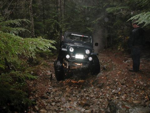 1985 Ford Bronco - A dark-colored Jeep CJ is navigating a rocky, wooded trail with a person standing to the right watching.