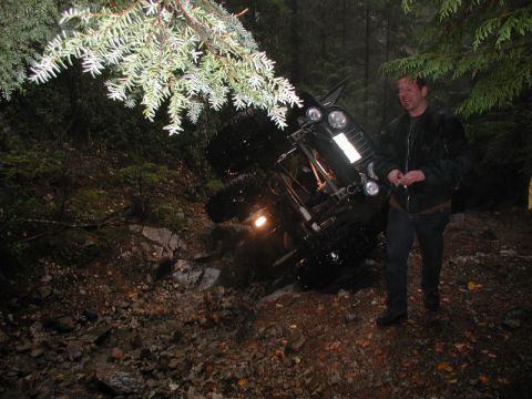 1985 Ford Bronco - A black Jeep Wrangler is overturned on a rocky, wooded trail. A man in a black jacket stands nearby, smiling.