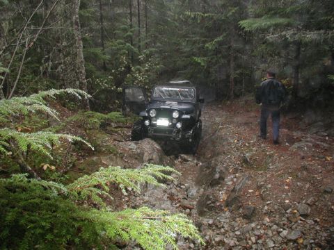 1985 Ford Bronco - A dark-colored Jeep CJ is navigating a rocky, muddy trail in a dense forest. A person in a dark jacket walks behind the vehicle.