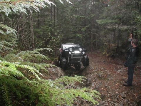 1985 Ford Bronco - A black Jeep Wrangler navigates a rocky, wooded trail with a person standing to the right, gesturing.