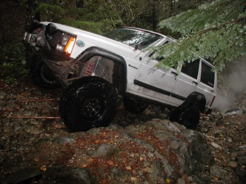 1985 Ford Bronco - A white Jeep Cherokee is navigating a rocky, wooded trail with large off-road tires. The vehicle is tilted at an angle as it climbs over ob…