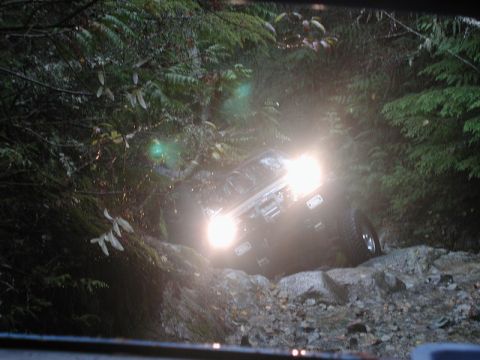 1985 Ford Bronco - A dark-colored SUV navigates a rocky, wooded trail with its headlights on. The vehicle is angled, showing its front and side as it climbs o…