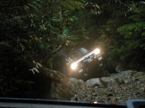 1985 Ford Bronco - A dark-colored SUV, possibly a 1985 Ford Bronco, navigates a rocky, wooded trail with its headlights on. The vehicle is tilted at an angle…