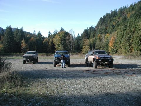 1985 Ford Bronco - Three lifted off-road trucks are parked in a gravel lot with a wooded hillside in the background. A person is standing between the two truc…