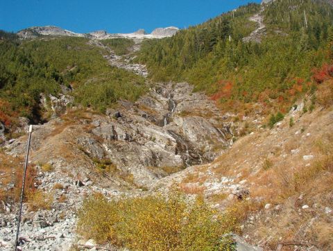 1985 Ford Bronco - A steep, rocky mountainside with scattered evergreen trees and some deciduous trees showing fall colors. A thin waterfall cascades down the…