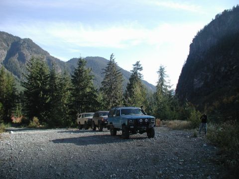 1985 Ford Bronco - A light blue 1980s Ford Bronco is parked on a gravel road in a mountainous, forested area. The vehicle is facing the camera at a three-quar…