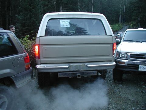 1985 Ford Bronco - The rear of a tan 1980s Ford Bronco is shown with a trailer hitch and exhaust smoke billowing from the tailpipe. A gray Jeep Grand Cherokee…