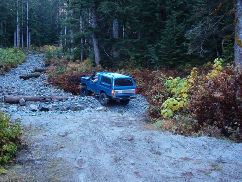 1985 Ford Bronco - A blue Ford Bronco is driving off-road on a rocky, wooded trail during the autumn season. The vehicle is navigating a steep incline with lo…