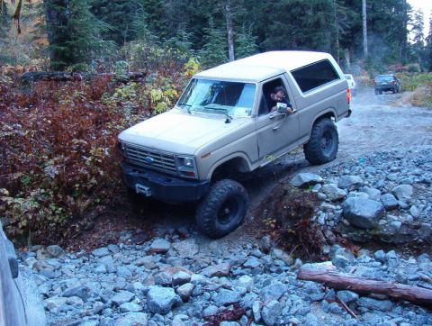 1985 Ford Bronco - A tan 1980s Ford Bronco is navigating a rocky, wooded trail. The vehicle is angled to the right, with its front tires on a higher elevation…