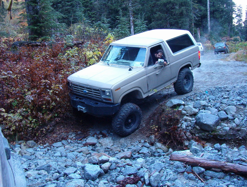 Tan 1980s Ford Bronco off-roading on a rocky trail in a forest.