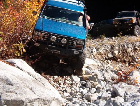 1985 Ford Bronco - A blue off-road van navigates a rocky, uneven trail with autumn foliage on the left. Another off-road vehicle is visible further up the tra…