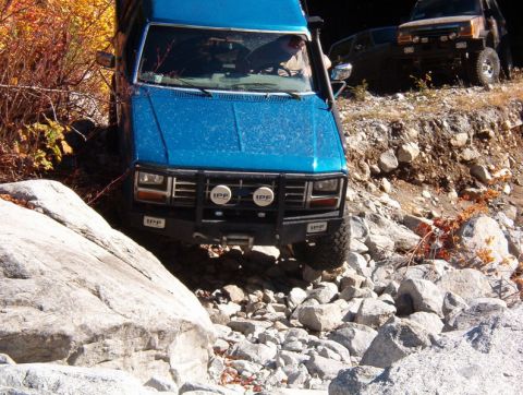 1985 Ford Bronco - A blue off-road SUV navigates a rocky trail with a second vehicle visible in the background. The front of the SUV features a black bumper w…