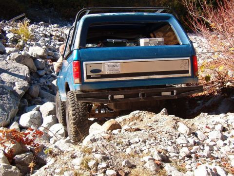 1985 Ford Bronco - The rear view of a teal Ford Bronco is shown navigating a rocky, off-road trail. The vehicle has large, aggressive tires and a custom rear…