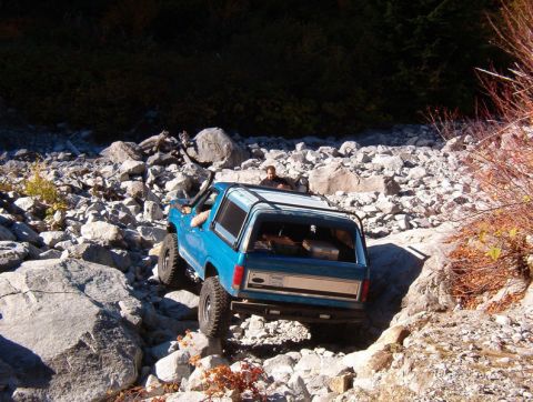 1985 Ford Bronco - A blue Ford Bronco is navigating a rocky, off-road trail. The vehicle is angled to the right, showing its rear and side profile as it climb…