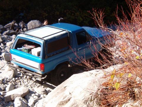 1985 Ford Bronco - A blue 1985 Ford Bronco is navigating a rocky, off-road terrain. The rear of the vehicle is visible, with gear stored in the open cargo are…