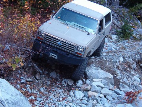 1985 Ford Bronco - A tan and white 1980s Ford Bronco is navigating a rocky, off-road trail with steep inclines and dense foliage. The vehicle is tilted to the…
